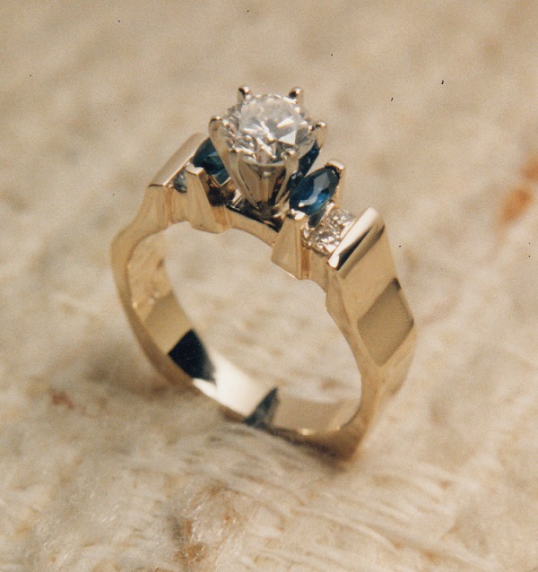 Gorgeous round brilliant diamond in an exquisite hand carved yellow gold mounting flanked by two marquis Sapphires which in turn are flanked by a total of four channel set round diamonds two on each side.